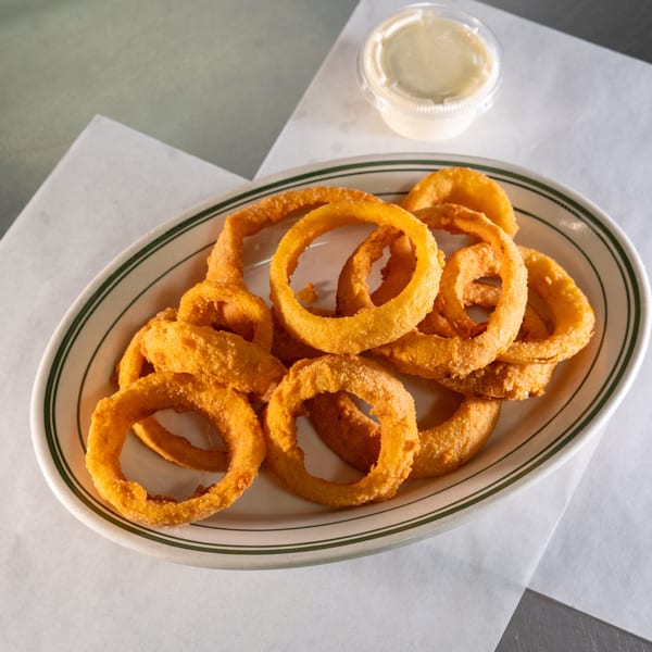 A plate of crispy, golden-brown onion rings with a side of dipping sauce on a white paper liner.