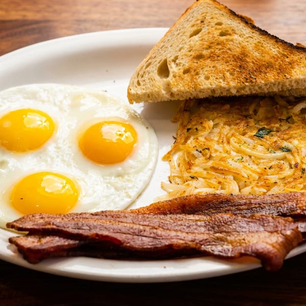 Breakfast plate with three fried eggs, hash browns, bacon, and toast on a wooden table.