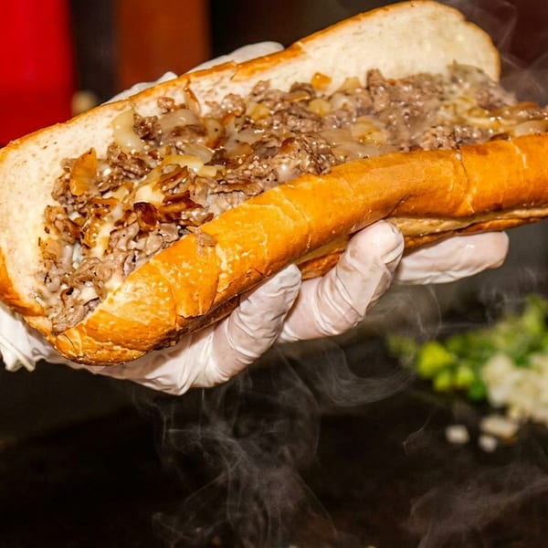 Steaming cheesesteak sandwich held by a gloved hand over a grill with chopped onions.