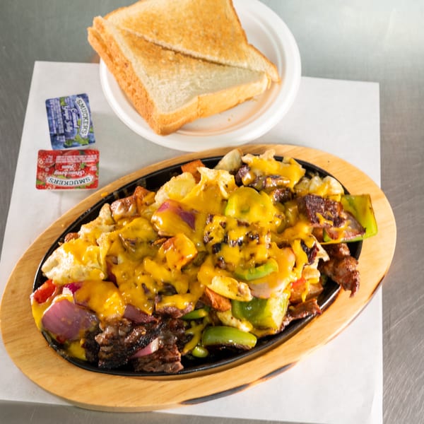 A sizzling skillet of steak, peppers, onions, and melted cheese, with toast and jam packets.