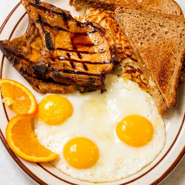 A breakfast plate with grilled pork chops, three fried eggs, hash browns, toast, and orange slices.