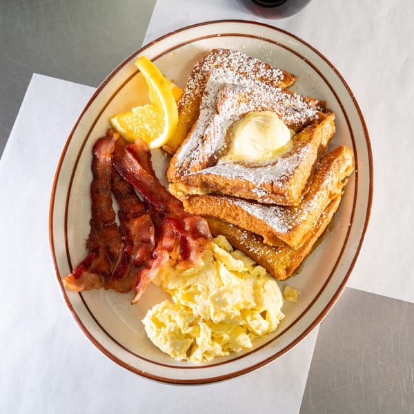 Breakfast plate with French toast, bacon, scrambled eggs, and an orange slice.