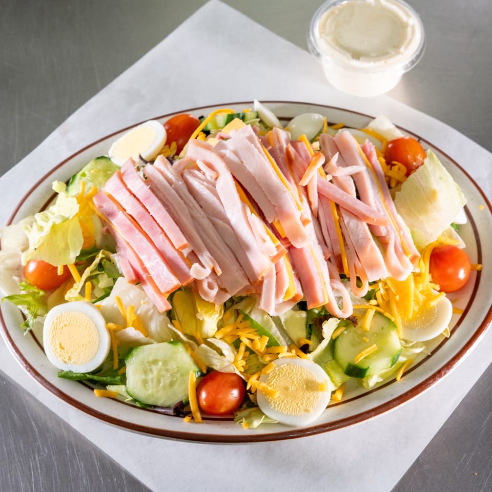 Chef salad with ham, cheese, hard-boiled eggs, tomatoes, and cucumbers on a white plate.